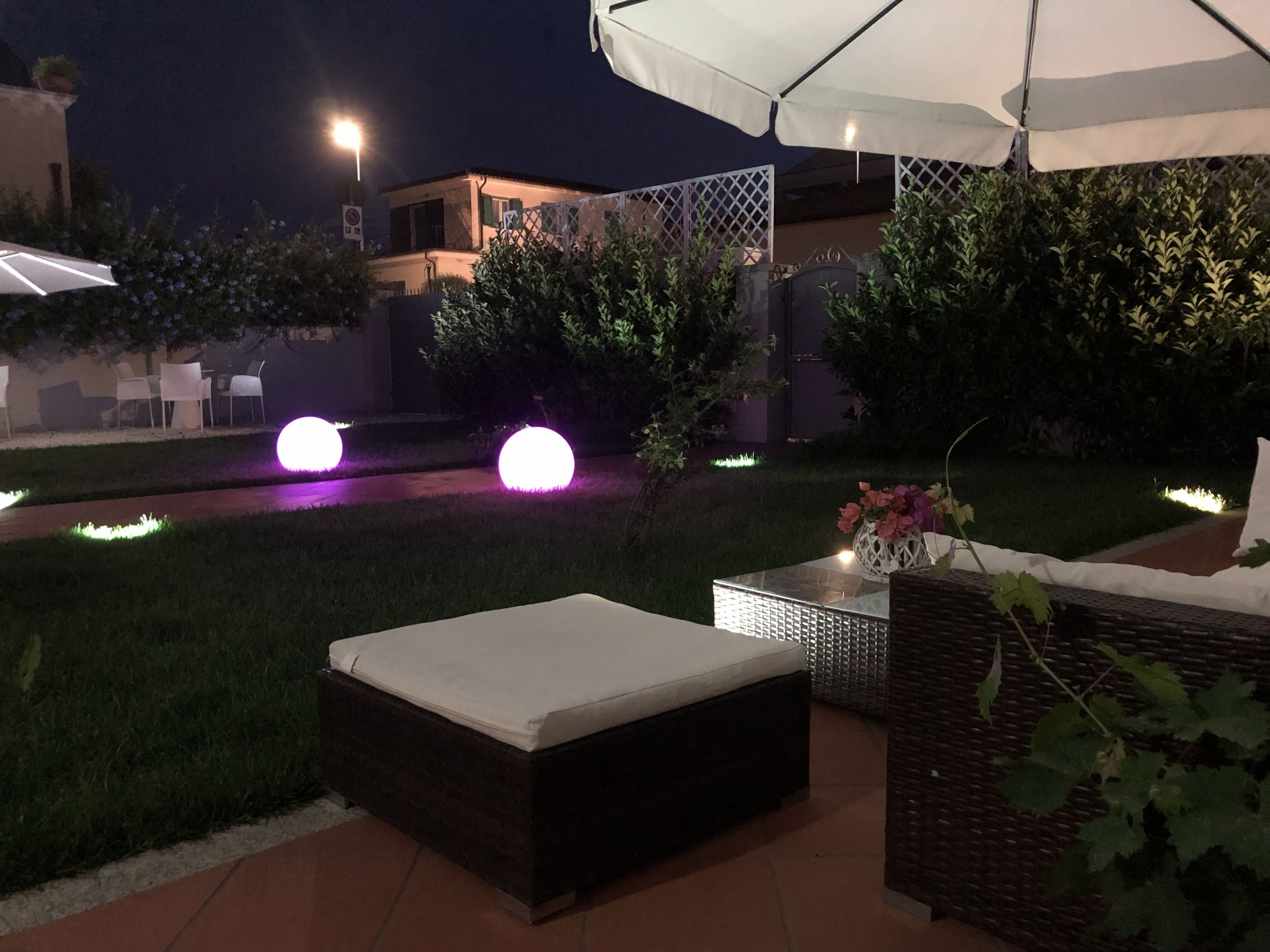 Un área de estar al aire libre con un sofá de mimbre y una mesa, rodeada de vegetación e iluminada con luces decorativas.