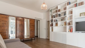 Ein modernes Wohnzimmer mit einer Couch, einem Holzschrank, weißen Regalen mit Büchern, einem Fernseher und einer Pendelleuchte.
