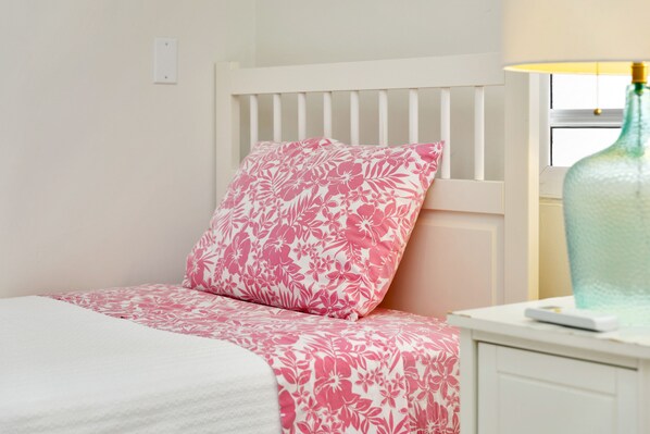 A bed with a white headboard and a pink floral pillow, a white bedside table with a lamp, and a window with blinds.