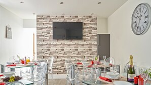 A modern dining area with glass tables and chairs, a brick-patterned wall, a mounted clock, and a flat-screen TV. - 88 Anfield (Liverpool)