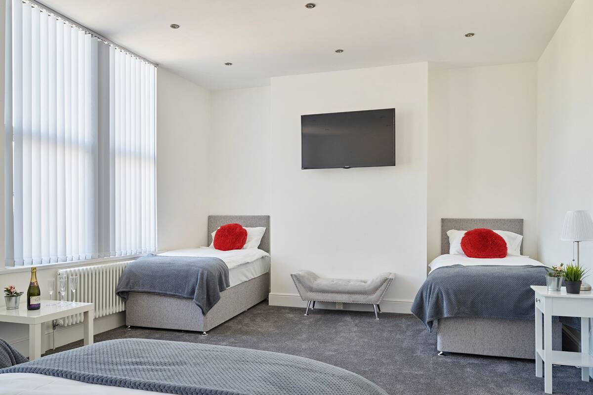A modern hotel room with two beds, a flat-screen TV, a small table with a lamp, and a radiator near the window.