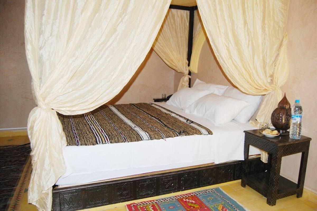Ein Bett mit Baldachin, ein Nachttisch mit einer Wasserflasche und ein gemustertes Teppich.