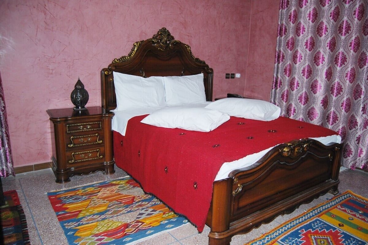 Ein Schlafzimmer mit einem hölzernen Bett, einem Nachttisch, einem gemusterten Teppich und rosafarbenen Wänden.