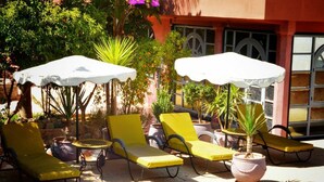 Zona junto a la piscina con sillas reclinables amarillas y sombrillas blancas, rodeada de plantas en macetas y un edificio con grandes ventanales.