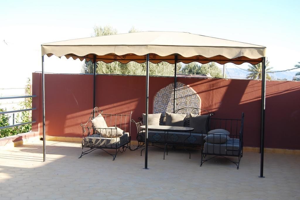 Een overdekt buitenterrein met metalen stoelen en een tafel, een behang met patroon en een rode muur op de achtergrond.