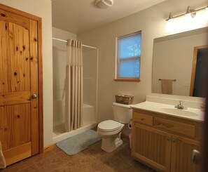 A bathroom with a wooden door, a shower with a beige curtain, a white toilet, a wooden vanity with a mirror, and a window with blinds. - Long Range Mountain & Valley Views from this Beautiful Chalet (Murphy)