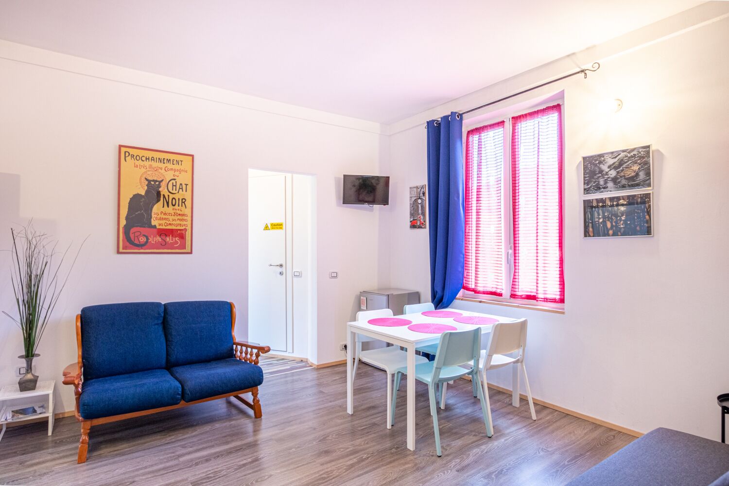 A room with a blue sofa, a white table with pink placemats, a TV on the wall, and a window with pink blinds.