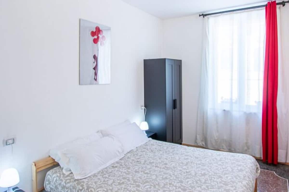 Un dormitorio con cama, mesita de noche, armario y ventana con cortinas.