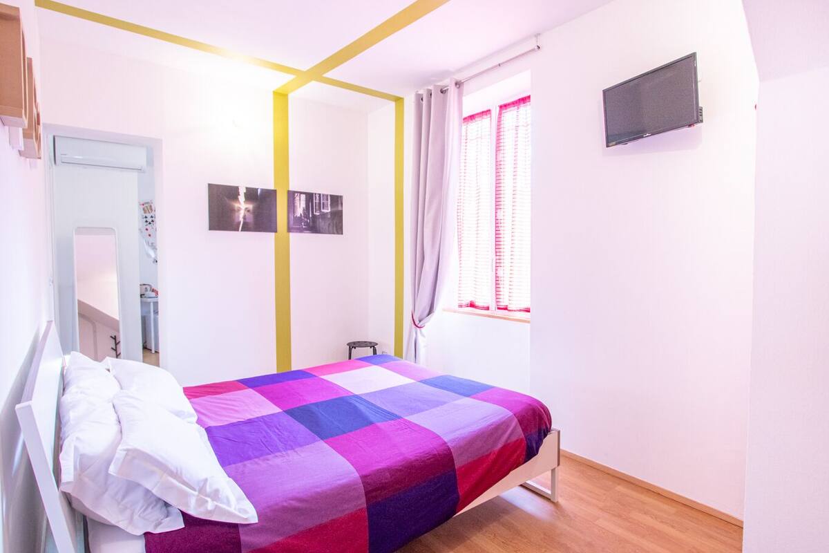 Un dormitorio con cama, televisor montado en la pared y ventana con cortinas.