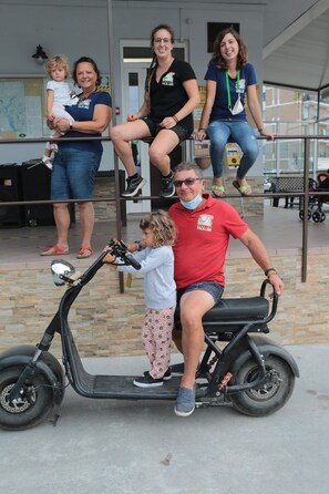 Une famille sur un scooter, un enfant vêtu d’un pull blanc et d’un pantalon rose, et des adultes en tenue décontractée.