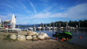 Un puerto deportivo junto al lago con barcos, una zona de playa con sillones de descanso y montañas al fondo.