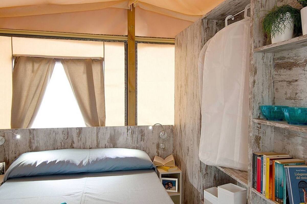 Un dormitorio con cama, ventana con cortinas, estantería con libros y repisa con objetos decorativos.
