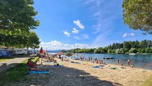 Una playa con tiendas de campaña y sombrillas, un lago y árboles.