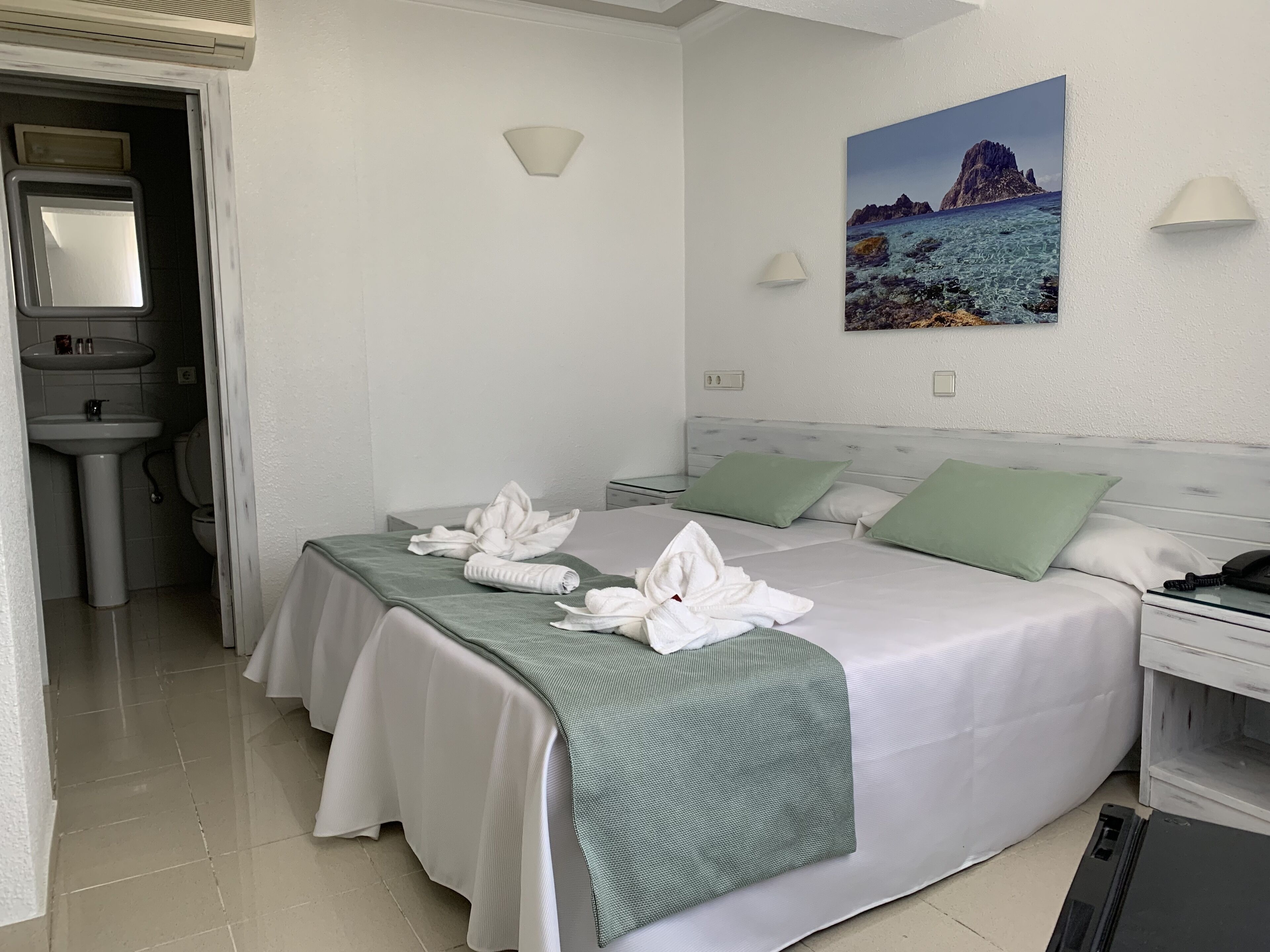 Habitación de hotel con cama, almohadas verdes, un cuadro de un paisaje costero, una mesita de noche y un baño con lavamanos e inodoro.