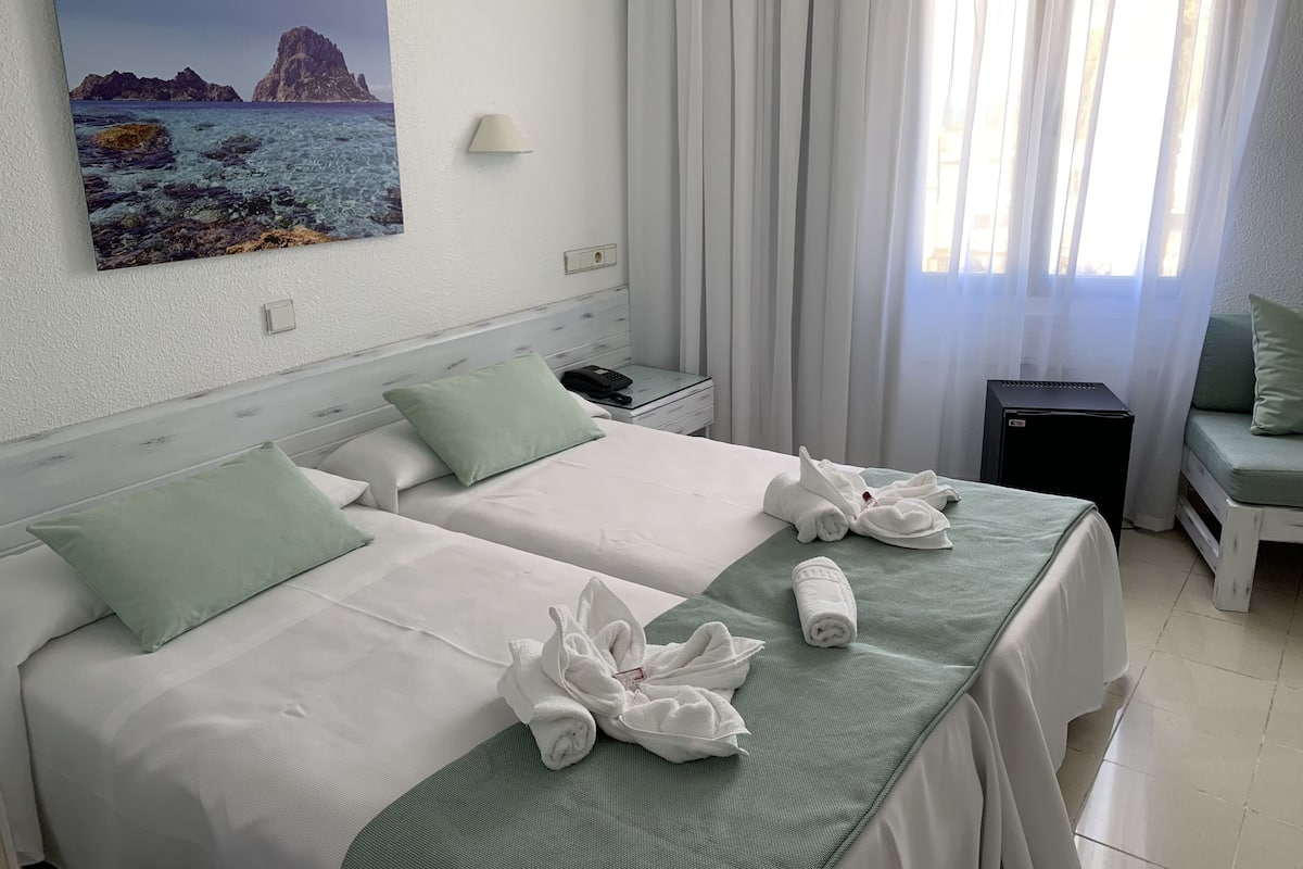 A hotel room with two beds, a painting of a coastal landscape, a lamp, a desk with a phone, and a small black speaker.