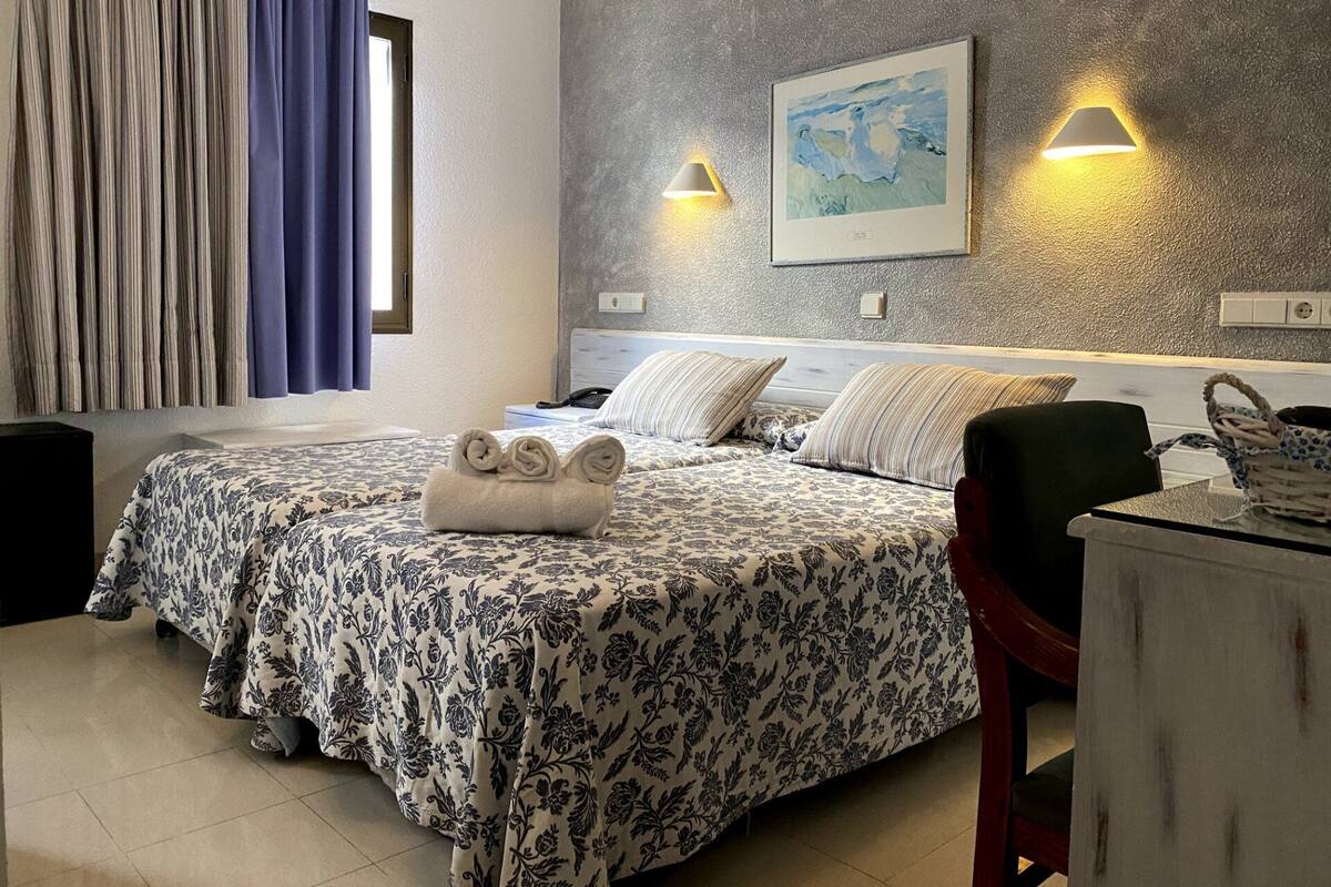 A hotel room with a bed, two bedside lamps, a painting on the wall, and a desk with a chair.