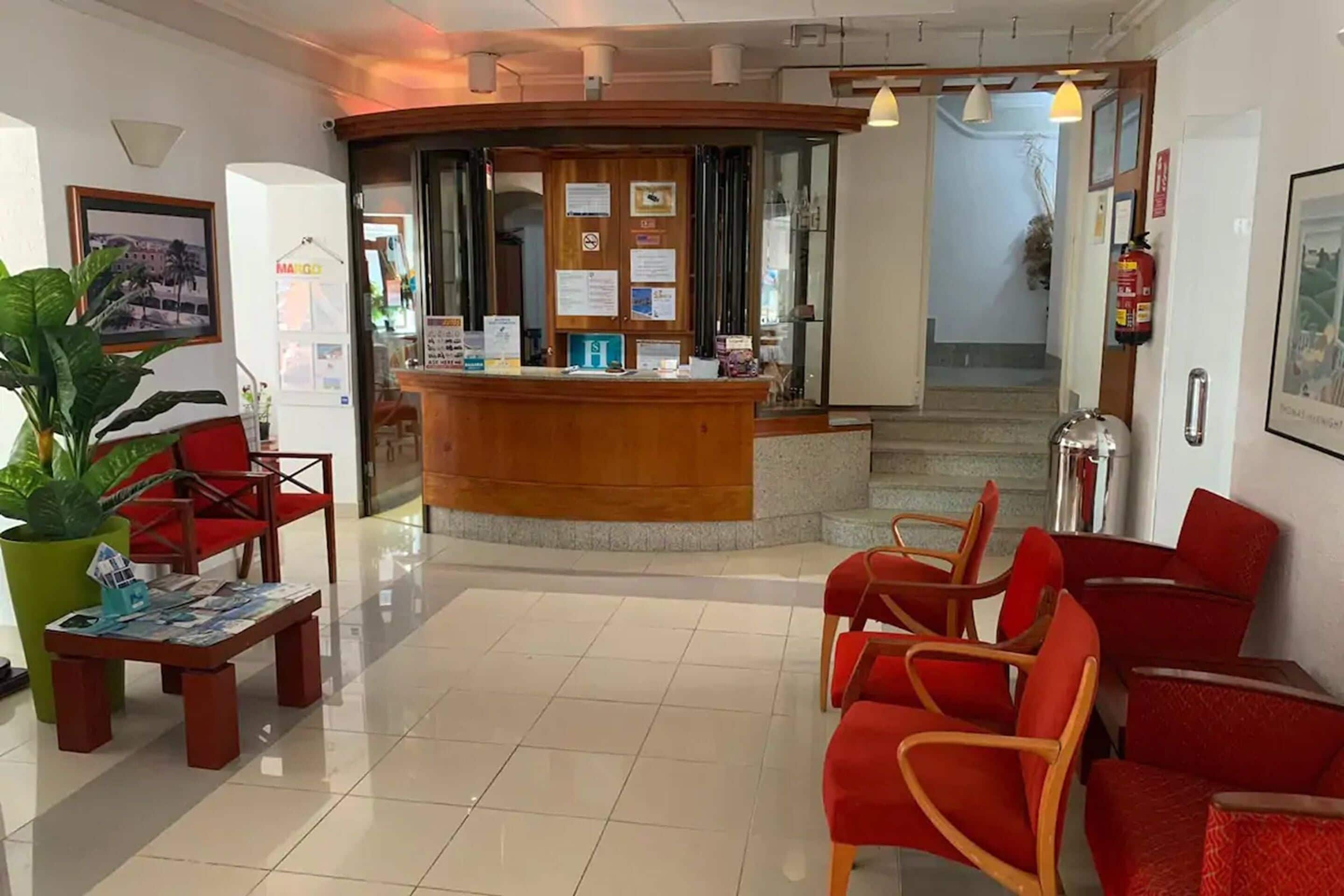 Zona de recepción de un hotel con un mostrador de madera, sillas rojas, una planta verde y suelo de baldosas.