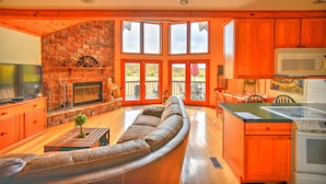 A spacious living area with a fireplace, wooden flooring, and a large window overlooking a deck.