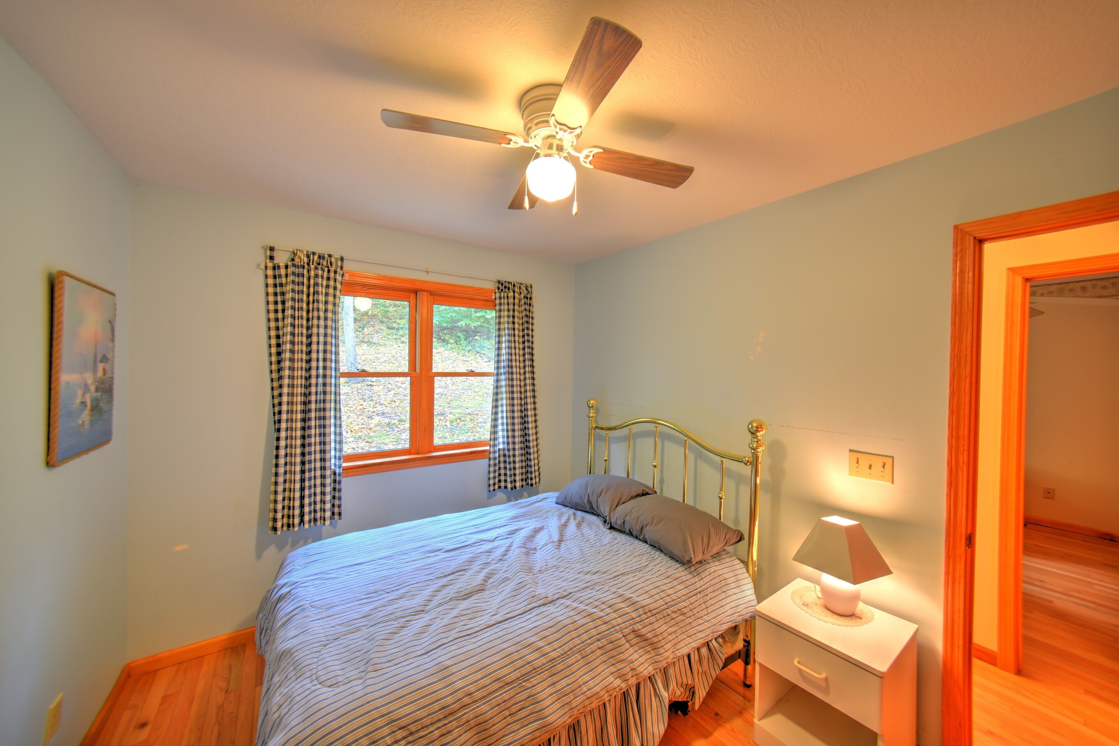 A bedroom with a bed, a nightstand with a lamp, a ceiling fan, a window with curtains, and a door.