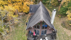 A cabin with a steeply pitched roof, a deck with outdoor seating, and a large window overlooking a forested area.