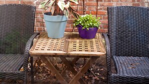 Een houten tafeltje met twee planten in potten en twee rieten stoelen, voor een bakstenen muur.