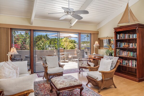 A spacious living room with wicker furniture, a large bookshelf, and a view of a tropical garden through sliding glass doors.