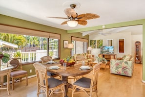 A spacious living room with a dining area, large windows, and a ceiling fan.