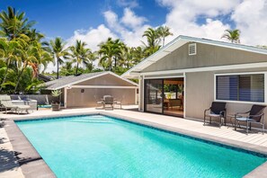 A modern pool area with clear blue water, surrounded by palm trees and a covered patio with outdoor furniture.