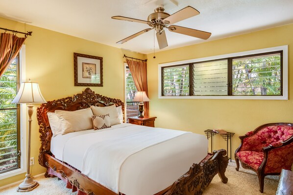 A bedroom with a large bed, a ceiling fan, a window with blinds, a red chair, and a nightstand with a lamp. - Beautiful, Tropical Vacation Rental in Keauhou! STVR-19-366304. (Kailua)