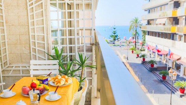 Un balcon avec une table dressée pour le petit-déjeuner, donnant sur une plage et une promenade bordée de plantes en pot et de parasols.