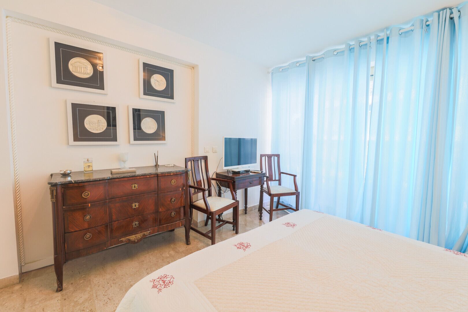 A bedroom with a bed, a dresser, two chairs, a desk with a computer, and a TV.