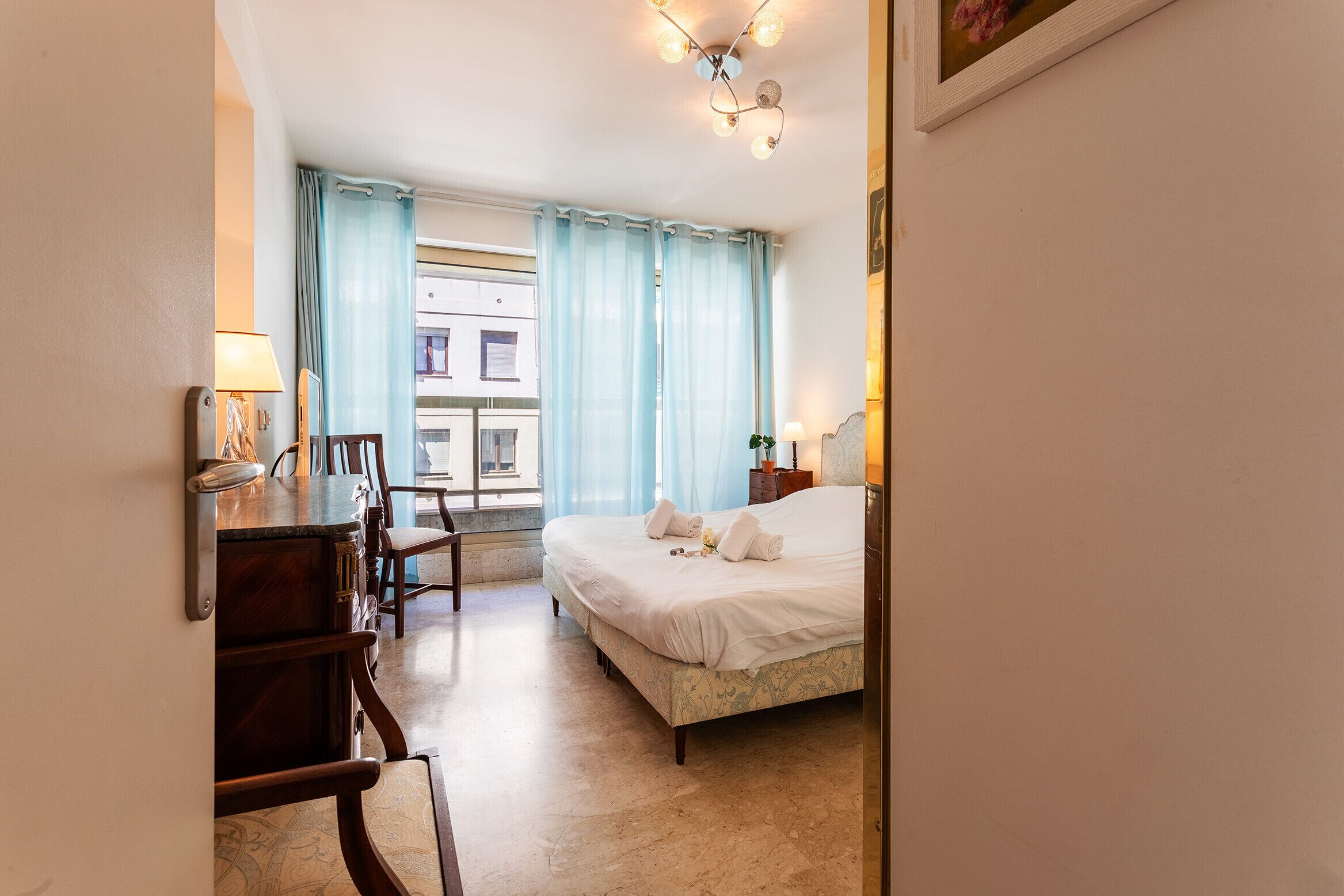 A hotel room with a bed, a desk with a lamp, a chair, and a window with curtains.