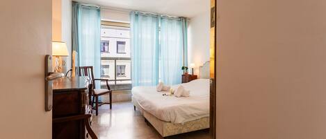 A hotel room with a bed, a desk with a lamp, a chair, and a window with curtains.