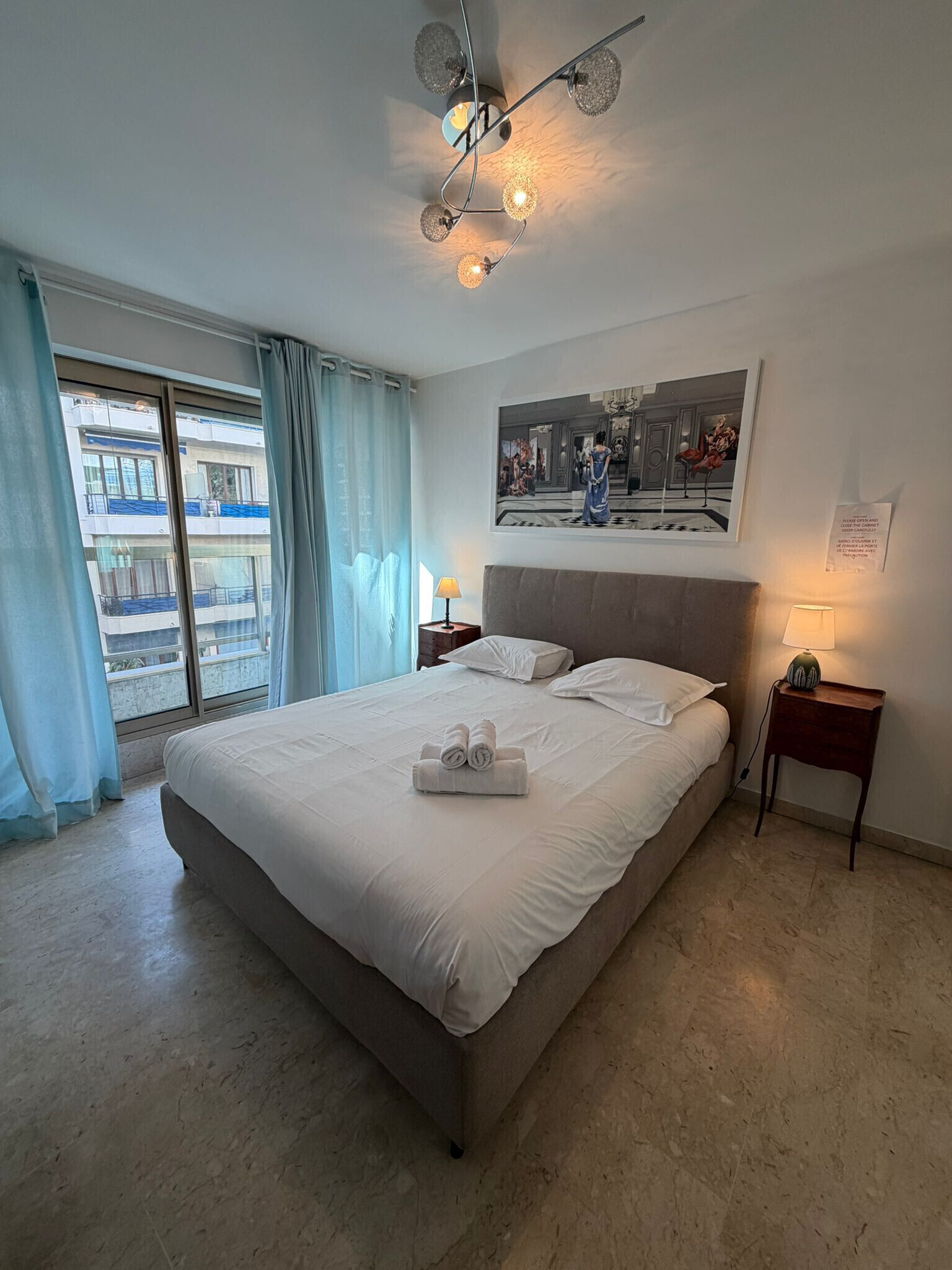 A hotel room with a large bed, a ceiling light fixture, a framed picture on the wall, and a view of the outside.