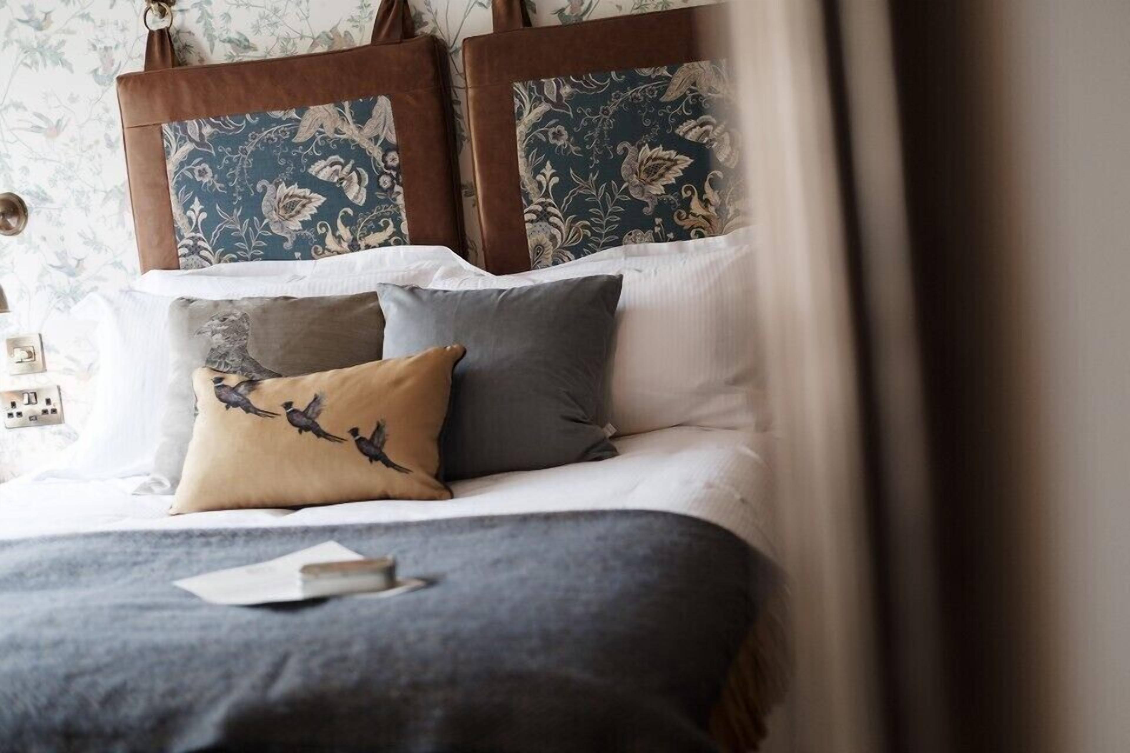 Een netjes opgemaakt bed met een blauwe deken, twee kussens met vogelmotieven en een hoofdbord met bloemenprint.