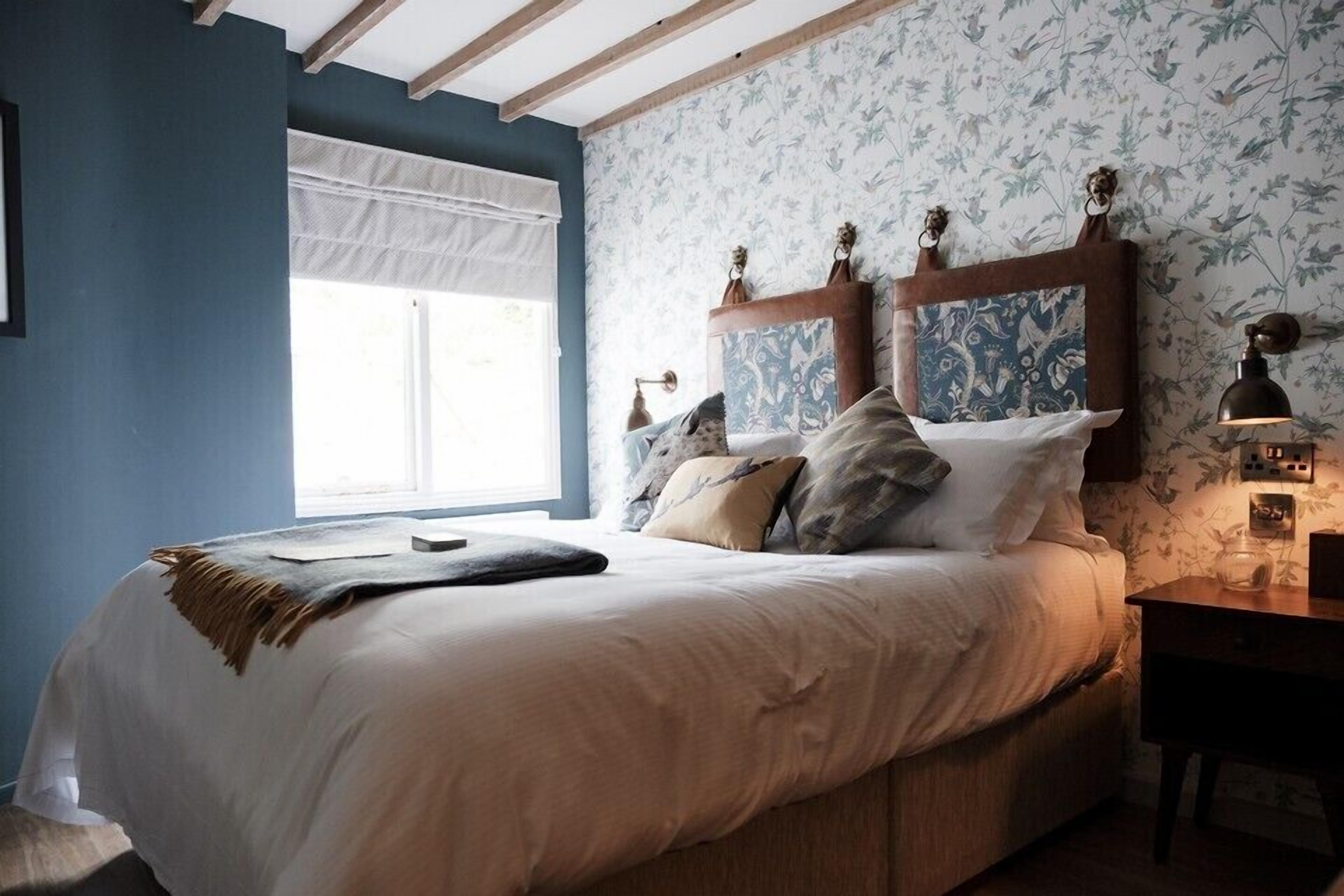 Ein Schlafzimmer mit einem Bett, einem Nachttisch, einer Lampe und einem Fenster mit weißem Vorhang.