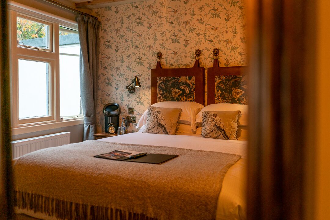 A bedroom with a double bed, floral wallpaper, a nightstand with a lamp, and a window with curtains.
