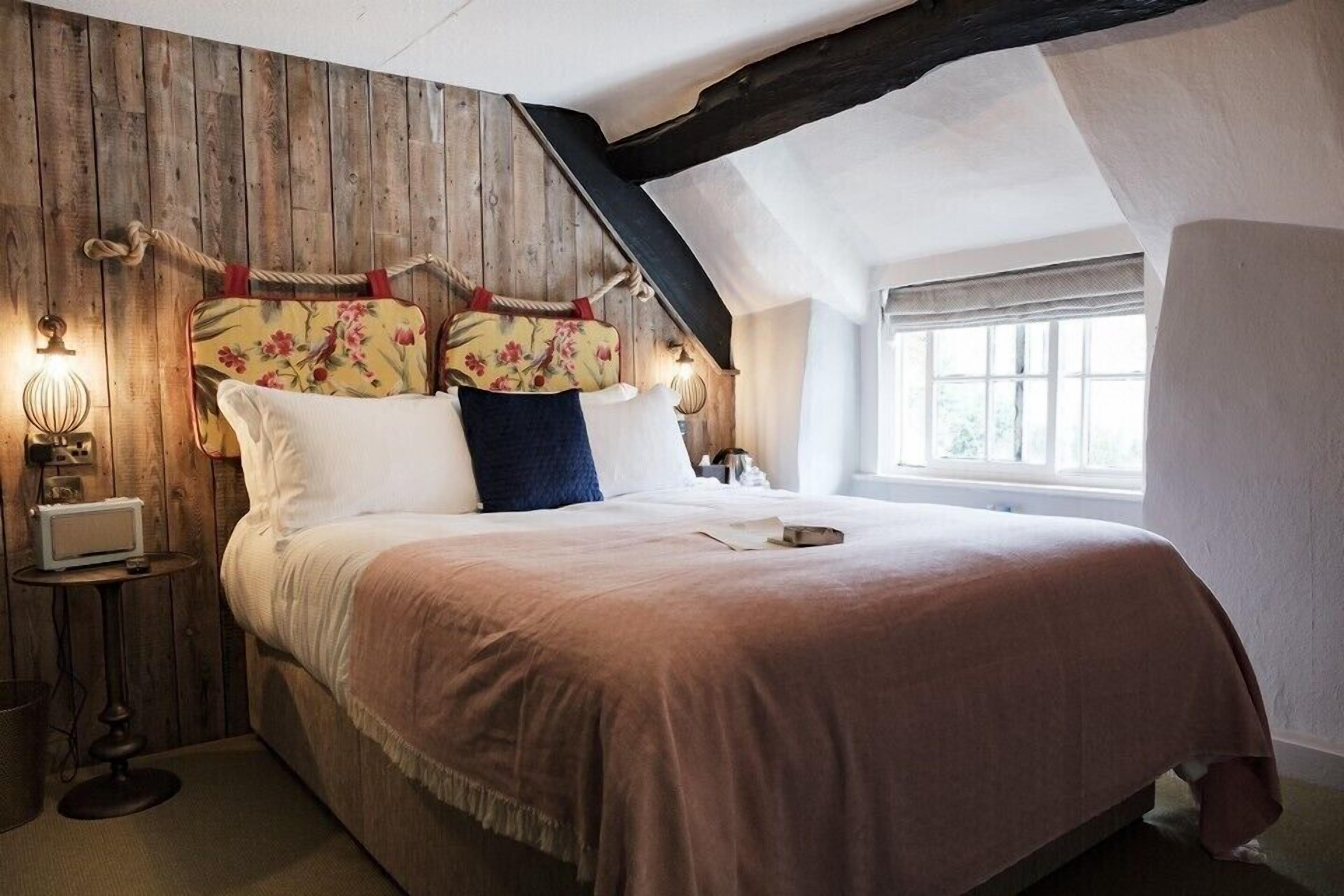 Ein Schlafzimmer mit einem Holzkopfteil, einem Bett mit weißer und rosa Bettwäsche, einem Nachttisch mit Lampe und einem Fenster mit Tageslicht.