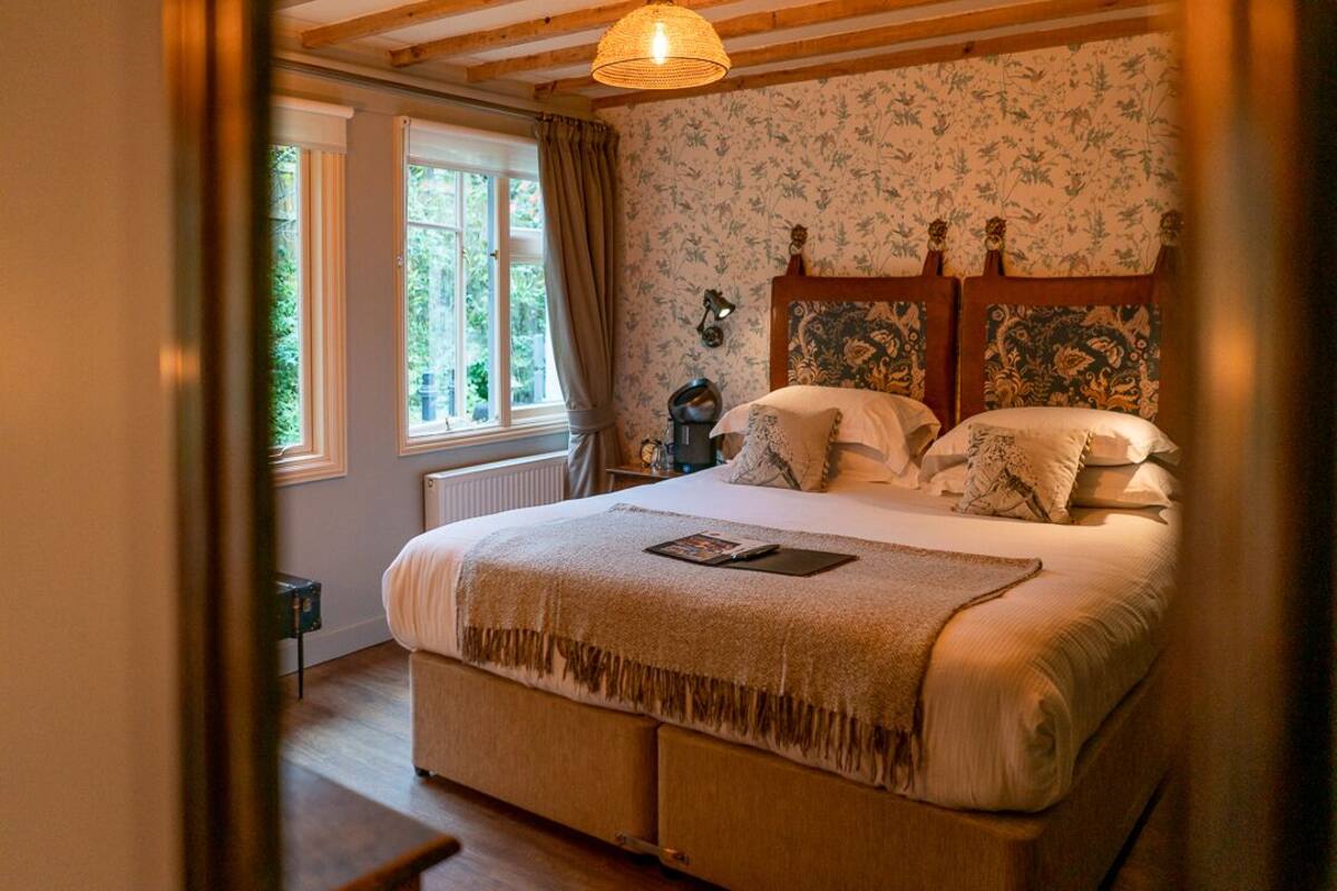 Une chambre avec un grand lit, une fenêtre avec des rideaux, un radiateur et un mur recouvert d’un papier peint à motifs floraux.