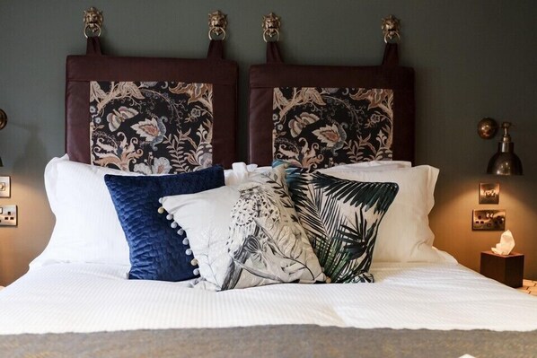 A neatly made bed with floral and tropical patterned pillows, a dark headboard, and a bedside table with a lamp and a tissue box. - Castle Inn (Wareham)
