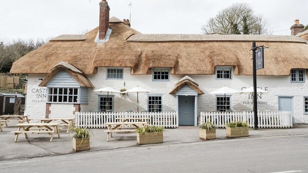 A traditional thatched-roof building with a white picket fence, labeled "CASTLE INN PUB & ROOMS," featuring outdoor seating and planters. - Castle Inn (Wareham)