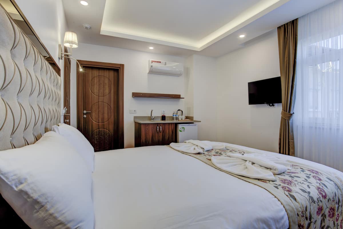 A hotel room with a large bed, a bedside table, a small desk with a mirror, a flat-screen TV mounted on the wall, and an air conditioning unit.