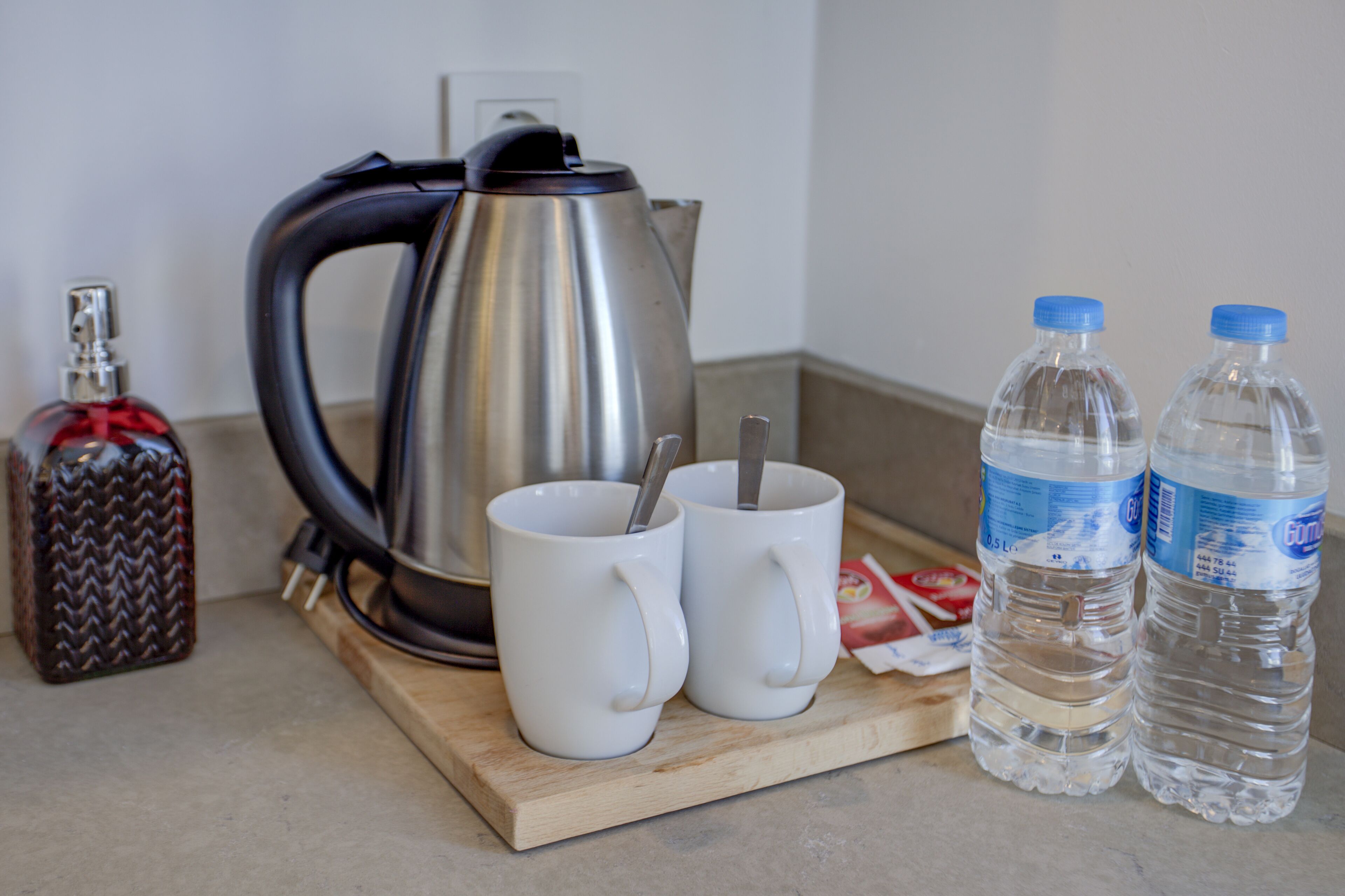 A kitchen counter with a stainless steel kettle, two white mugs with spoons, a bottle of water, and a decorative bottle.