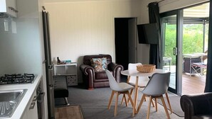 A modern kitchen with a gas stove and sink, a dining area with a table and chairs, a leather sofa, and a sliding glass door leading to an outdoor area. - New modern compact home, warm & cosy, Blue Bay beach bach! (Mahia)