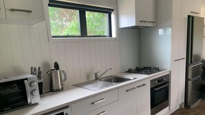 A modern kitchen with white cabinets, a stainless steel range, a built-in oven, a microwave, a sink, and a window with a view of greenery. - New modern compact home, warm & cosy, Blue Bay beach bach! (Mahia)