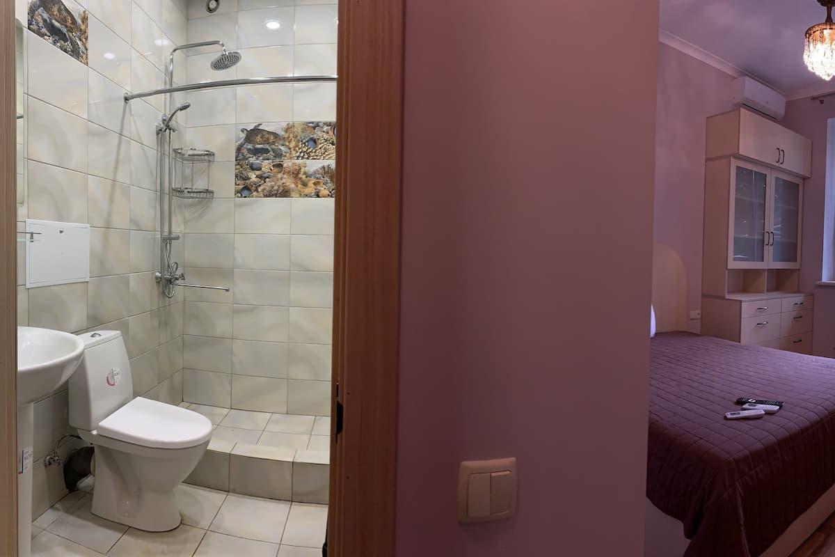 A bathroom with a shower, toilet, and sink. There is a bed with a blanket and a chair. A window provides natural light.
