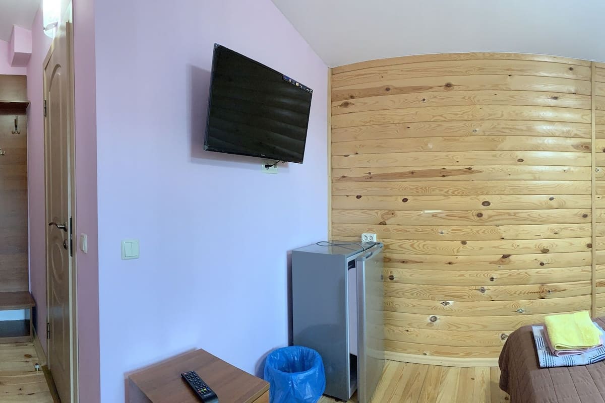 A room with a curved wooden wall, a bed with a patterned pillow, a small table with a remote control, and a trash bin, with a window providing a view of greenery outside.