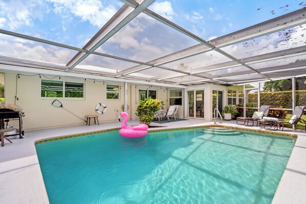 A covered swimming pool with a pink flamingo float, a grill, and a patio area with chairs and a table.