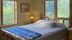 Una cama con estructura de madera, colchón acolchado y cabecera con barandilla. Hay una mesita de noche con una lámpara, un cuadro enmarcado en la pared y ventanas con vistas a la vegetación.