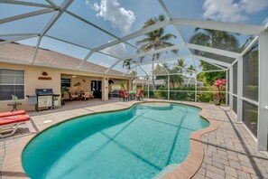 A covered outdoor swimming pool with a patio area, a barbecue setup, and a view of palm trees.